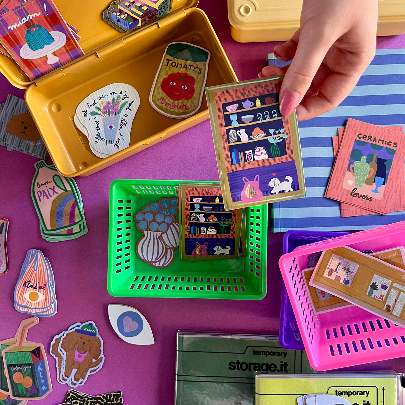 A hand holds a decorative sticker above colorful baskets filled with various stickers on a vibrant purple surface.