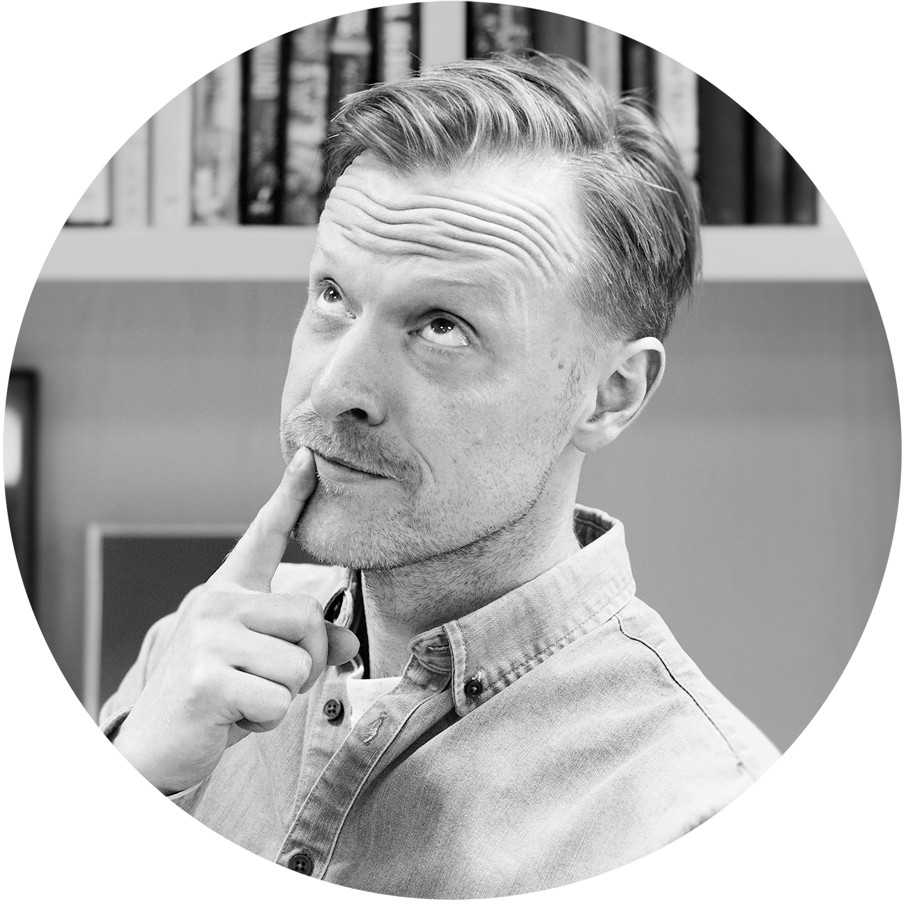 Black-and-white portrait of a man looking upward thoughtfully with his hand resting on his chin, seated indoors with a blurred bookshelf in the background.