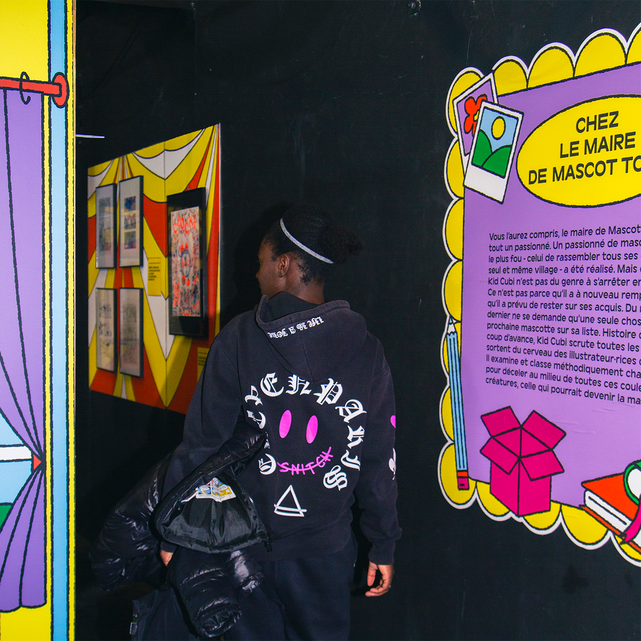 Person in a black hoodie stands near colorful, illustrated wall stickers with French text, in a vibrant, playful setting.