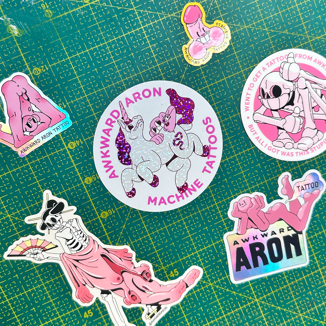 Die-cut stickers from tattoo artist Awkward Aron, printed on glitter, holographic and vinyl, set on a  cutting mat background.