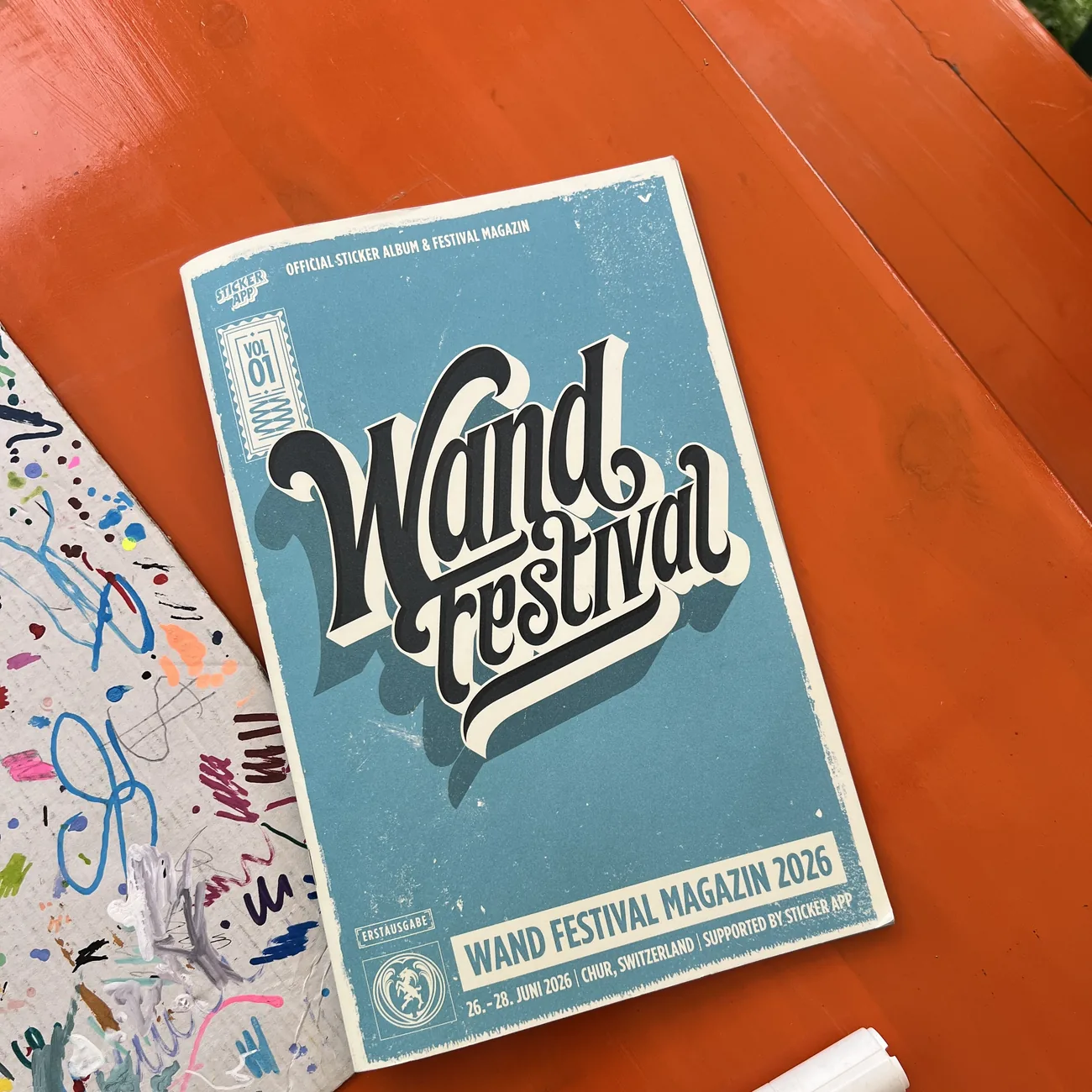 Oficjalny album z naklejkami i magazyn Wand Festival 2026 wspierany przez StickerApp obok markera z farbą na pomarańczowym stole