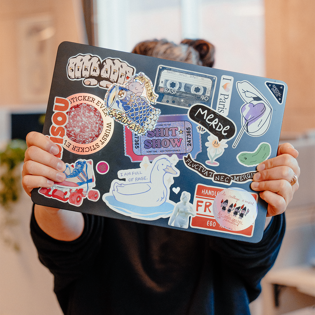 Close-up of a laptop covered in a mix of colorful stickers, including illustrated characters, typography, and graphic designs, held up by a person indoors.