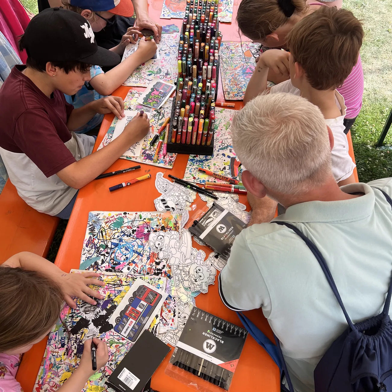Dzieci kolorują naklejki wycinane do kształtu Brainfart wokół stojaka z markerami z farbą podczas plenerowych warsztatów na festiwalu
