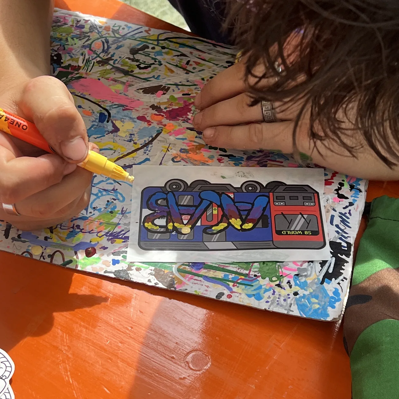 Dłoń koloruje etykietę z pociągiem S8 World żółtym markerem z farbą podczas warsztatów Brainfart x StickerApp