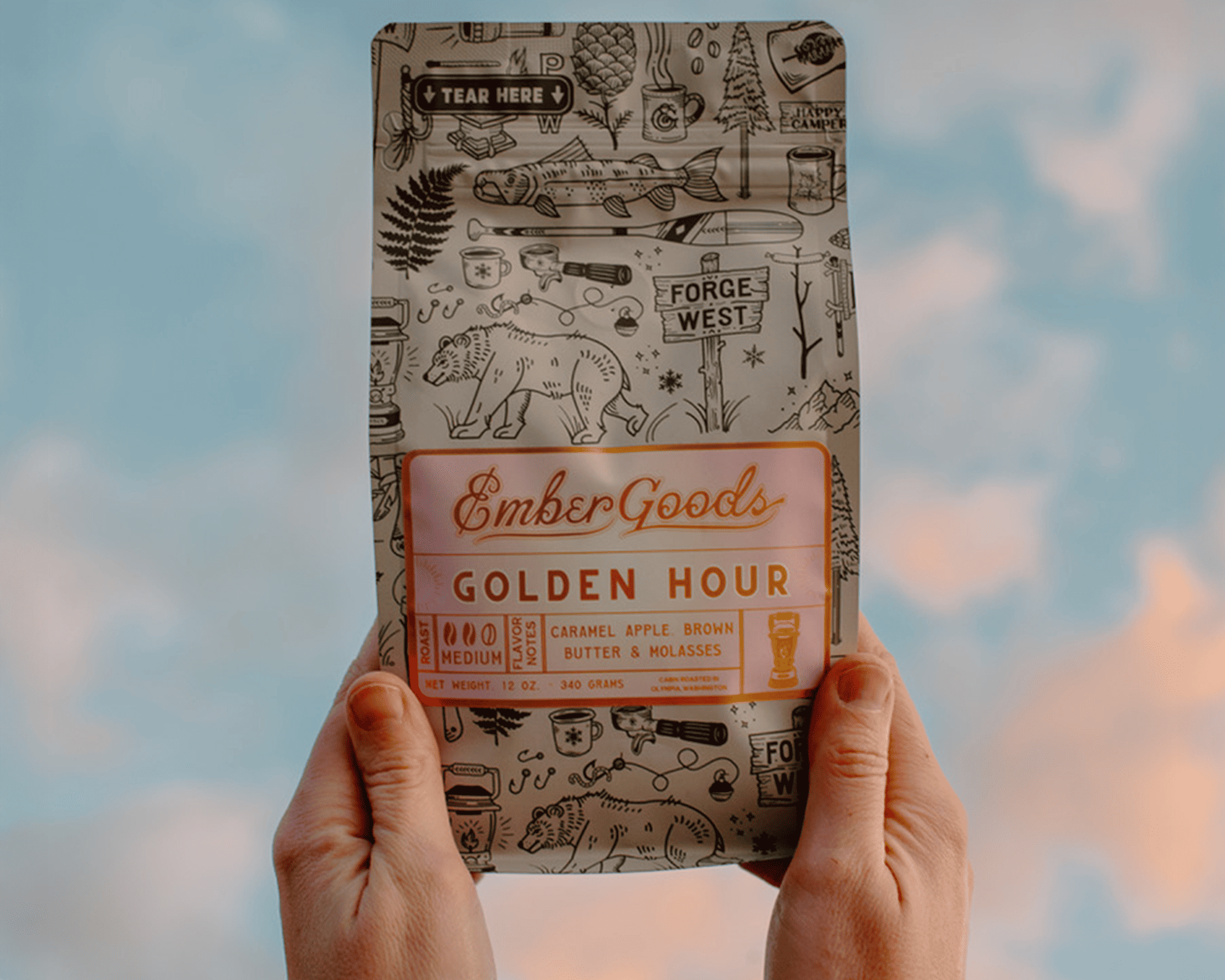 Dłonie trzymające torebkę kawy Ember Goods Golden Hour z metaliczną etykietą na tle nieba