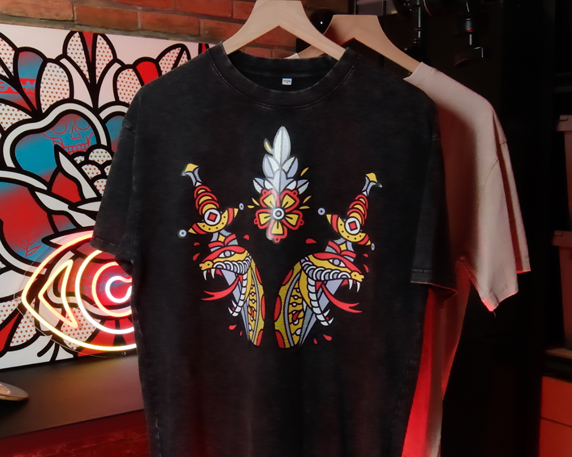 Black T-shirt with a colorful, symmetrical tribal design hangs in front of a beige shirt. A vibrant, abstract artwork is in the background.